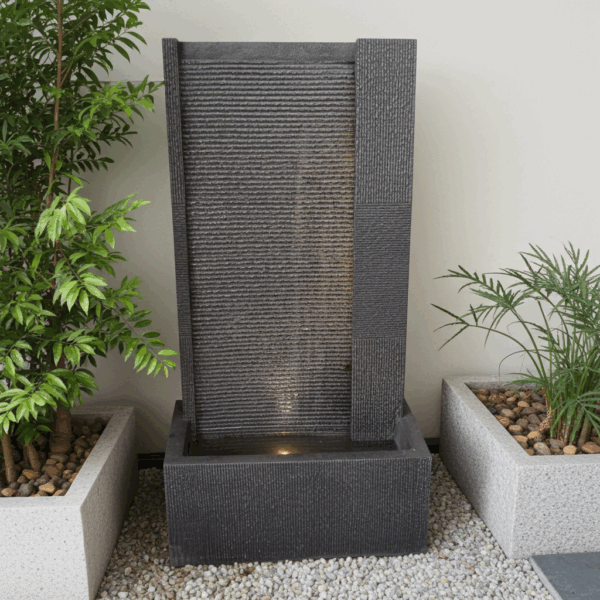 Fontaine cascade d'extérieur moderne de couleur anthracite avec éclairage LED. Mur d'eau rectangulaire en polyrésine à structure rainurée, placé entre deux bacs à plantes sur gravier clair.